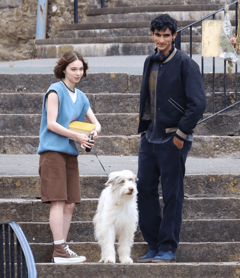 Teen girl with short hair walking dog and holding a box, talking to a tall Indian boy dressed in dark clothes.