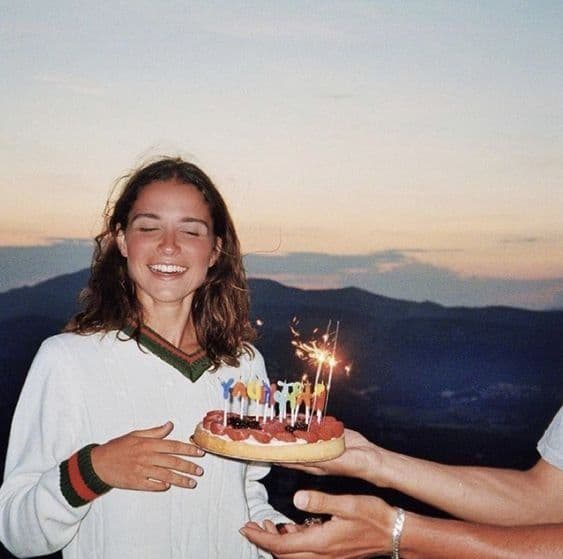 girl in front of mountain laughing with someone extending a birthday cake with sparklers in front of her