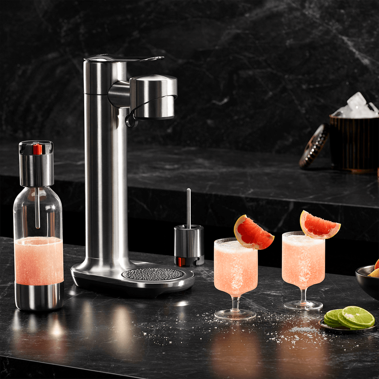 Stainless steel carbonator on black marble countertop with two bright peach cocktails in garnished glasses beside it.