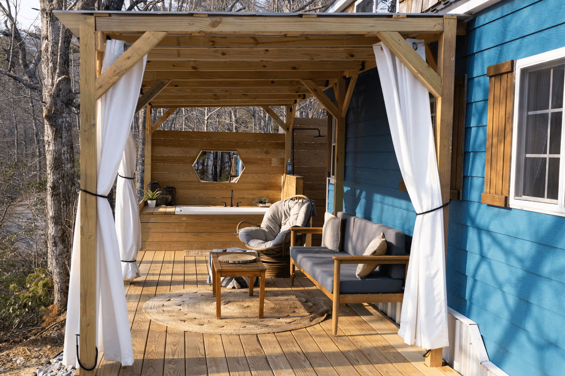 Outdoor patio showing a tub, seating, and privacy curtains in front of a blue mountainside cabin.