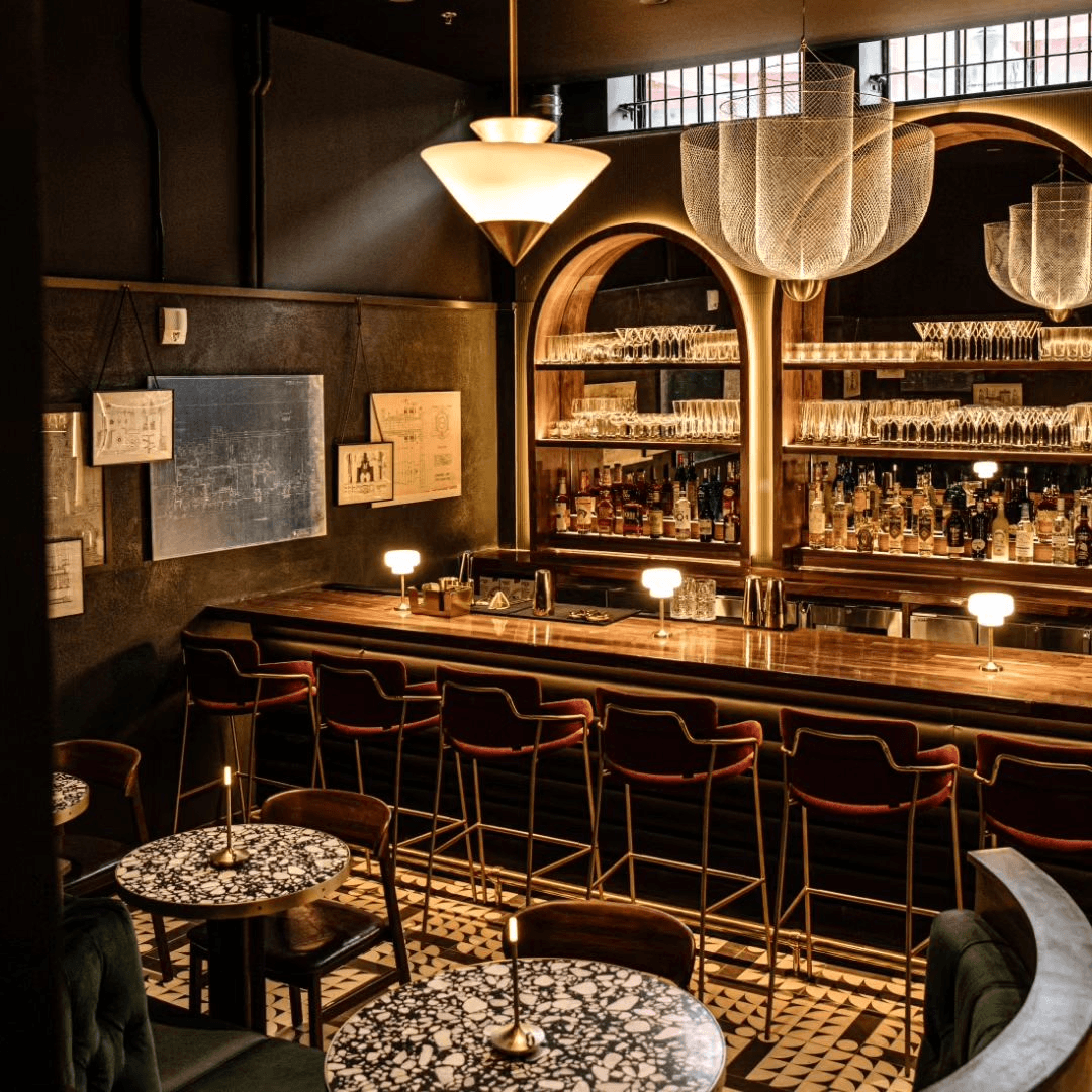 Moody speakeasy photographed at an angle. There's a gleaming bar with velvet stools, marble tables, and Art Deco lighting.
